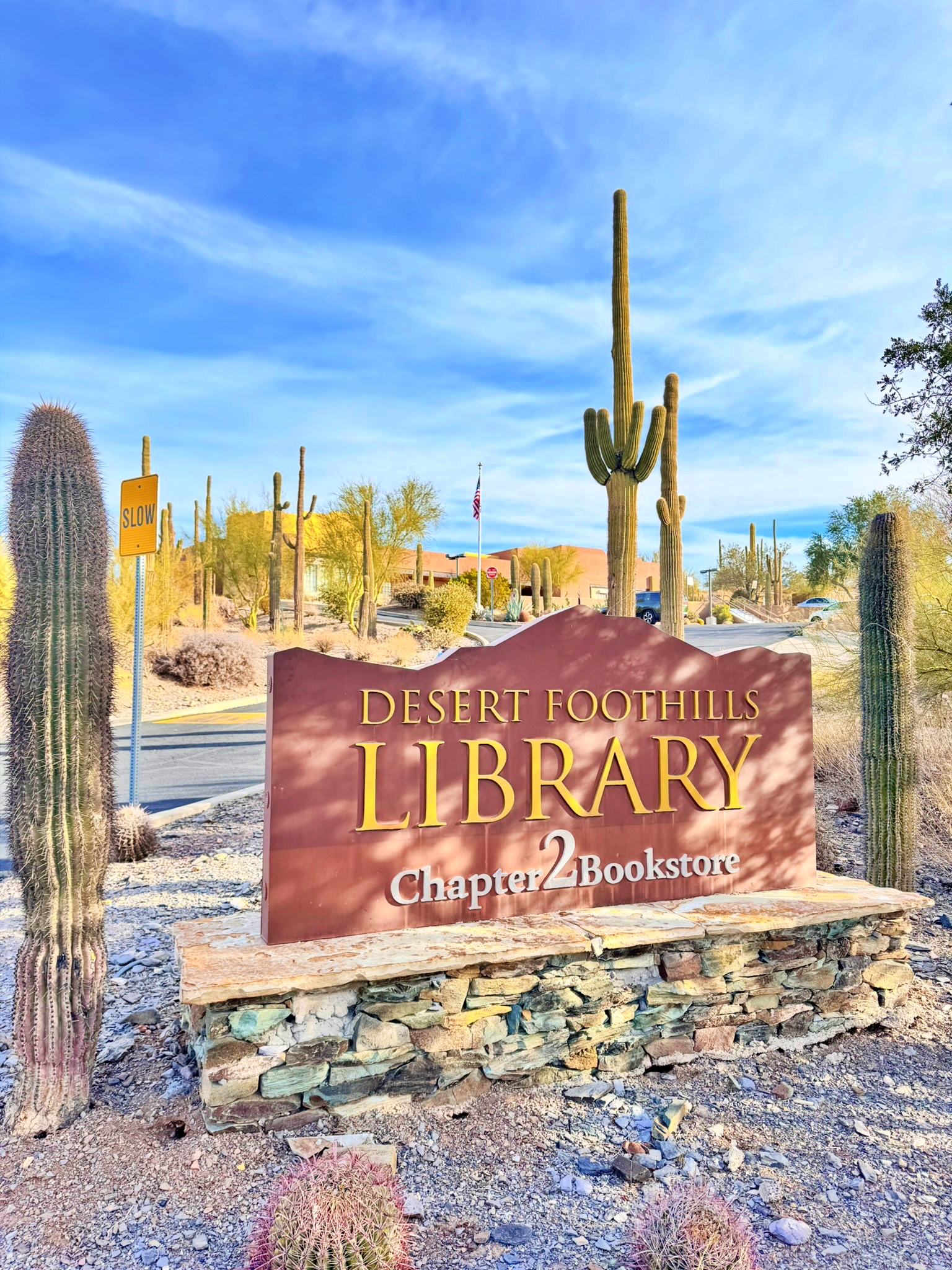 Entrance sign to the Desert Foothills Library in Cave Creek Arizona with the building and saguaros behind it.