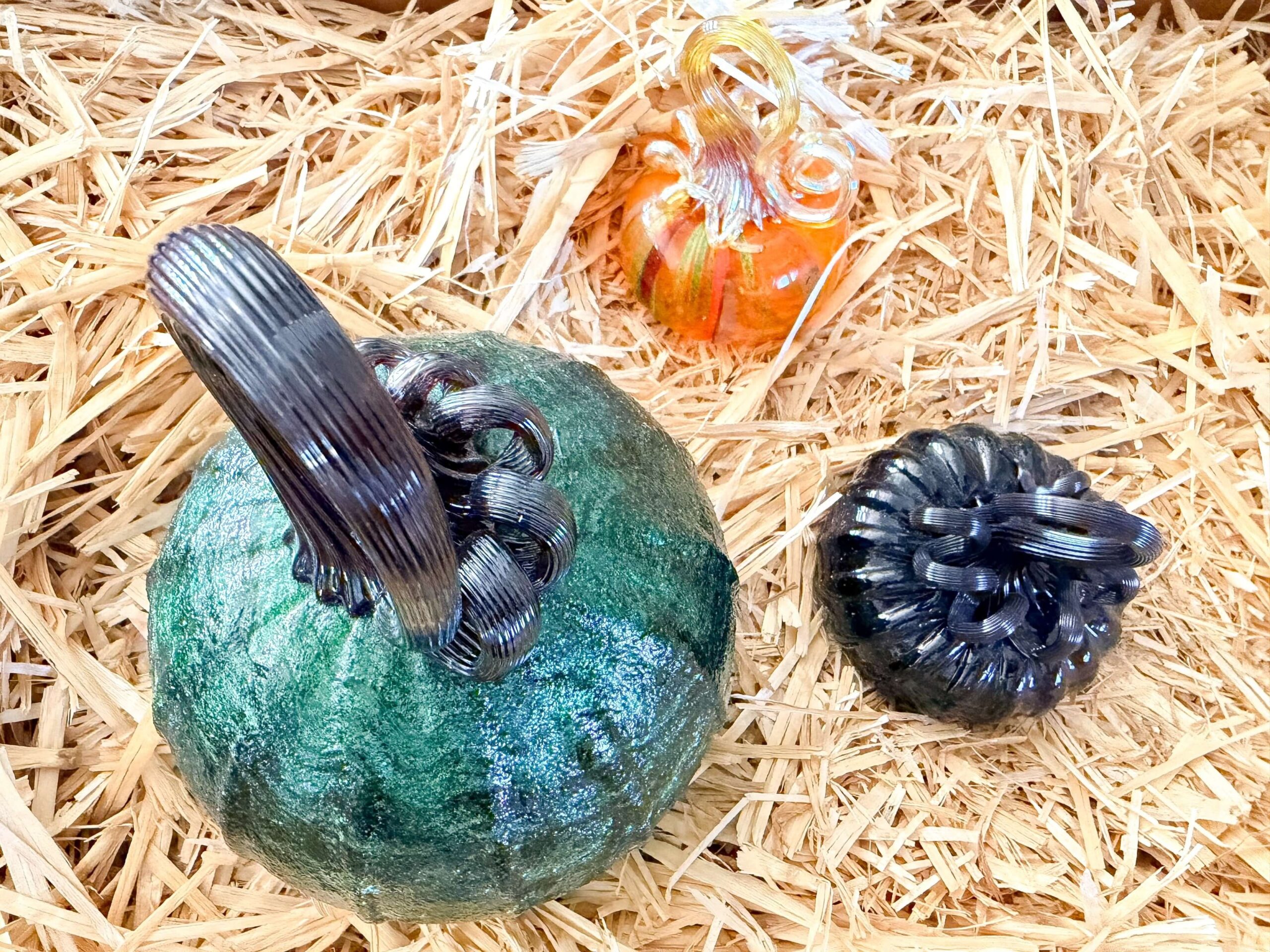 Close-up of colorful hand-blown glass pumpkins in green, black, and orange displayed on straw at the Glass Pumpkin Patch in Carefree, Arizona.