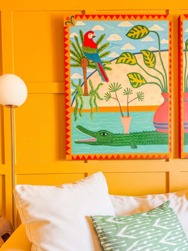 Golden yellow board and batten wall in a Cave Creek home with a folk art painting of a red parrot and green alligator, a white globe floor lamp, and a linen sofa with a green ikat pillow