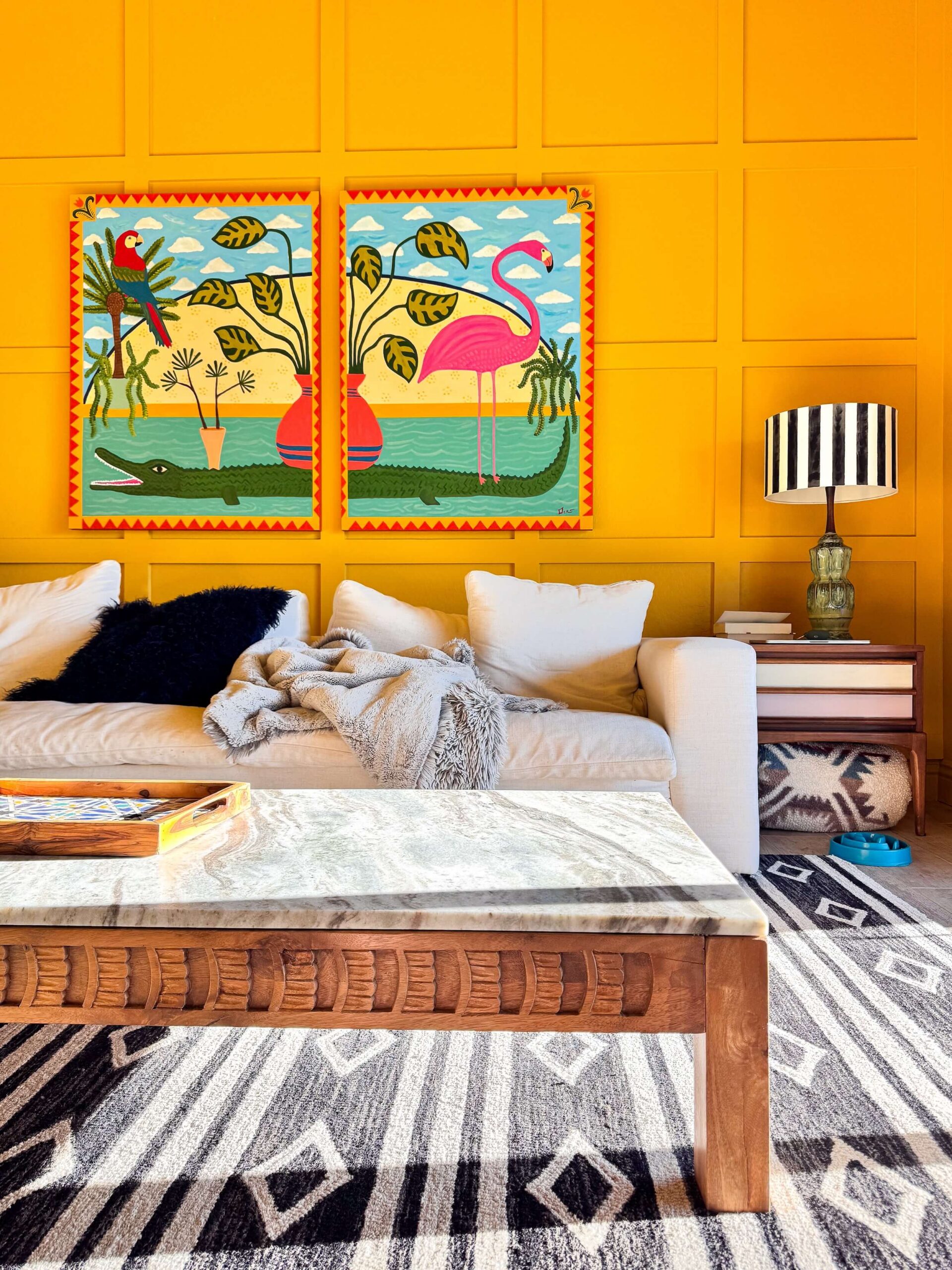 The Yellow Room in a Cave Creek home, golden yellow paneled walls with a two panel folk art painting of a flamingo, parrot and alligator above a white sofa, carved wood and marble coffee table, and black and white diamond rug